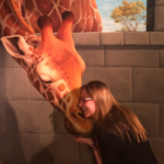 A foto mostra uma mulher branca na frente de uma parede pintada que mostra uma girafa com o pescoço inclinado para baixo. Amy parece estar abraçando o focinho da girafa. Ela tem cabelo loiro e liso e está usando um suéter preto e óculos.
