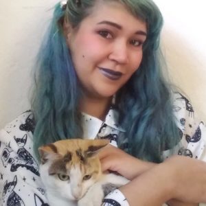 A foto quadrada mostra uma mulher branca com cabelo azul na altura do peito e um batom azulado. Está com uma camisa branca de botões com uma estampa preta de gatinhos, e no colo segura um gato tricolor. Está sorrindo para a câmera, com a cabeça levemente tombada para o lado